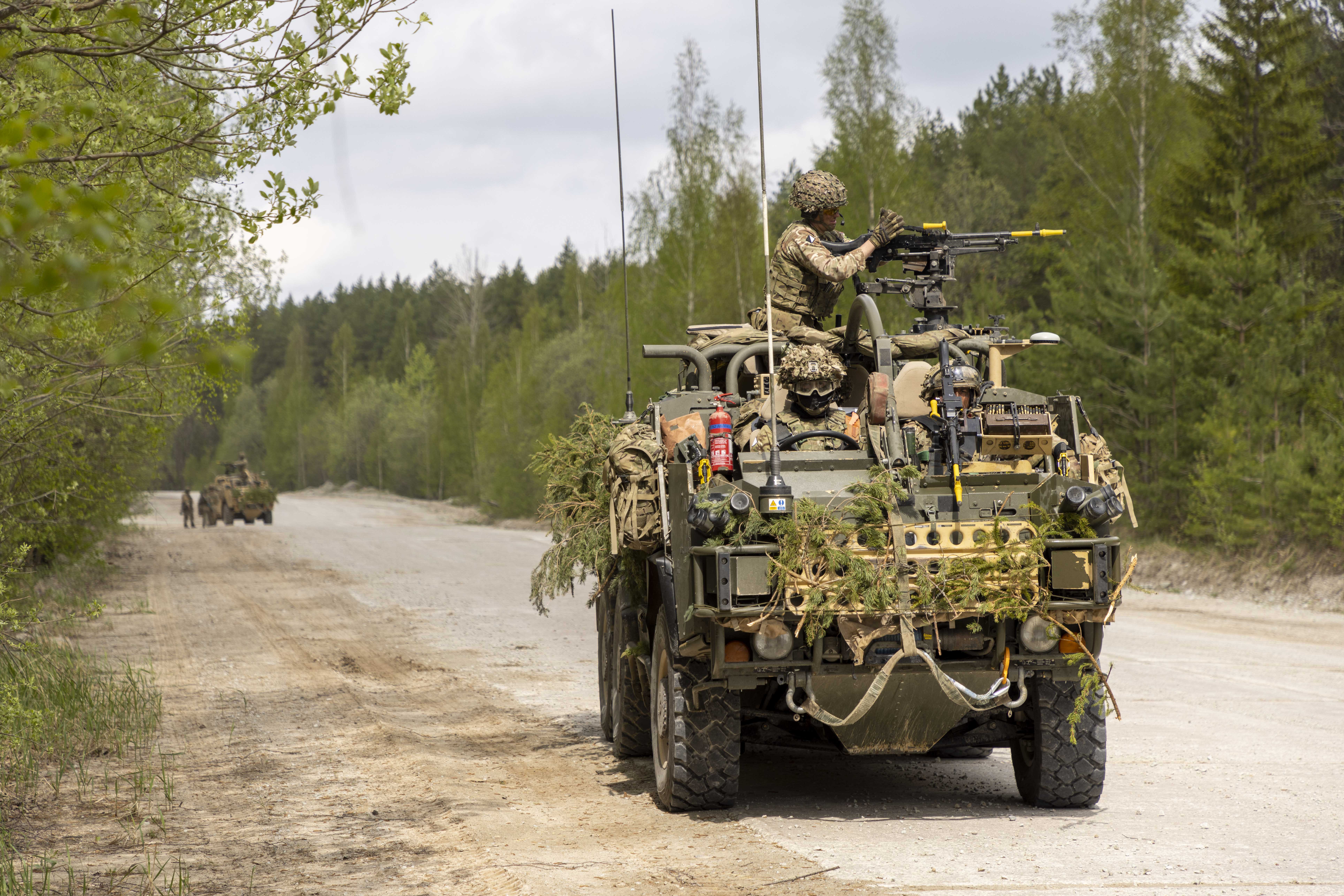 A military vehicle equipped with camouflage and a mounted machine gun is parked on a dirt road in a forested area. 
