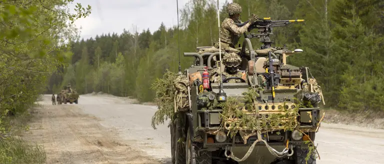 A military vehicle equipped with camouflage and a mounted machine gun is parked on a dirt road in a forested area.