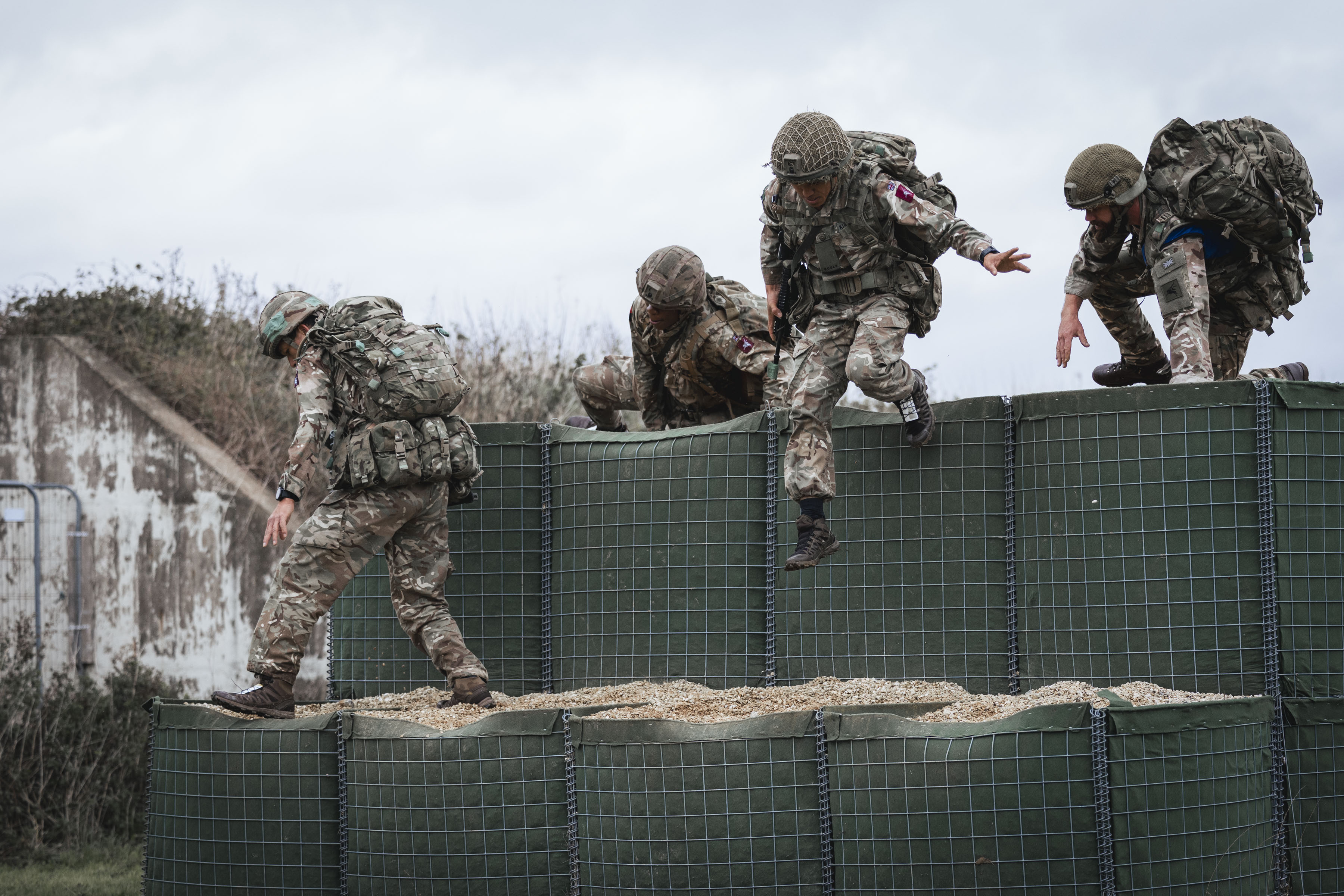 Four soldiers in full camouflage gear scale an obstacle during a training exercise.