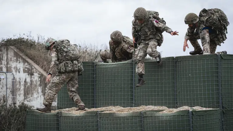 Four soldiers in full camouflage gear scale an obstacle during a training exercise.