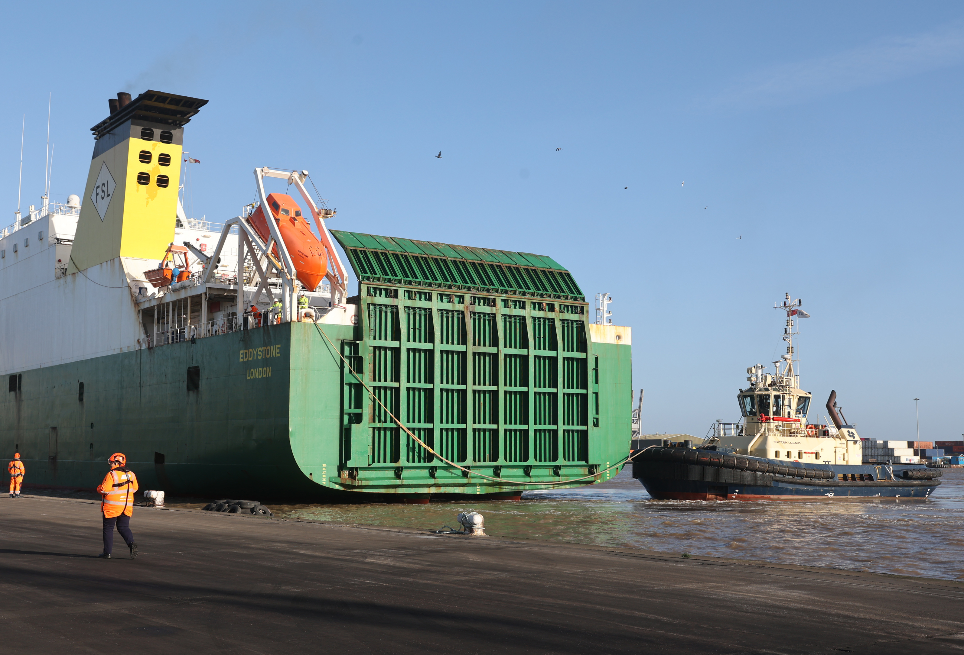 A large green roll on, roll of ferry is moored in a port. A tug boat is nudging the side of the ferry.