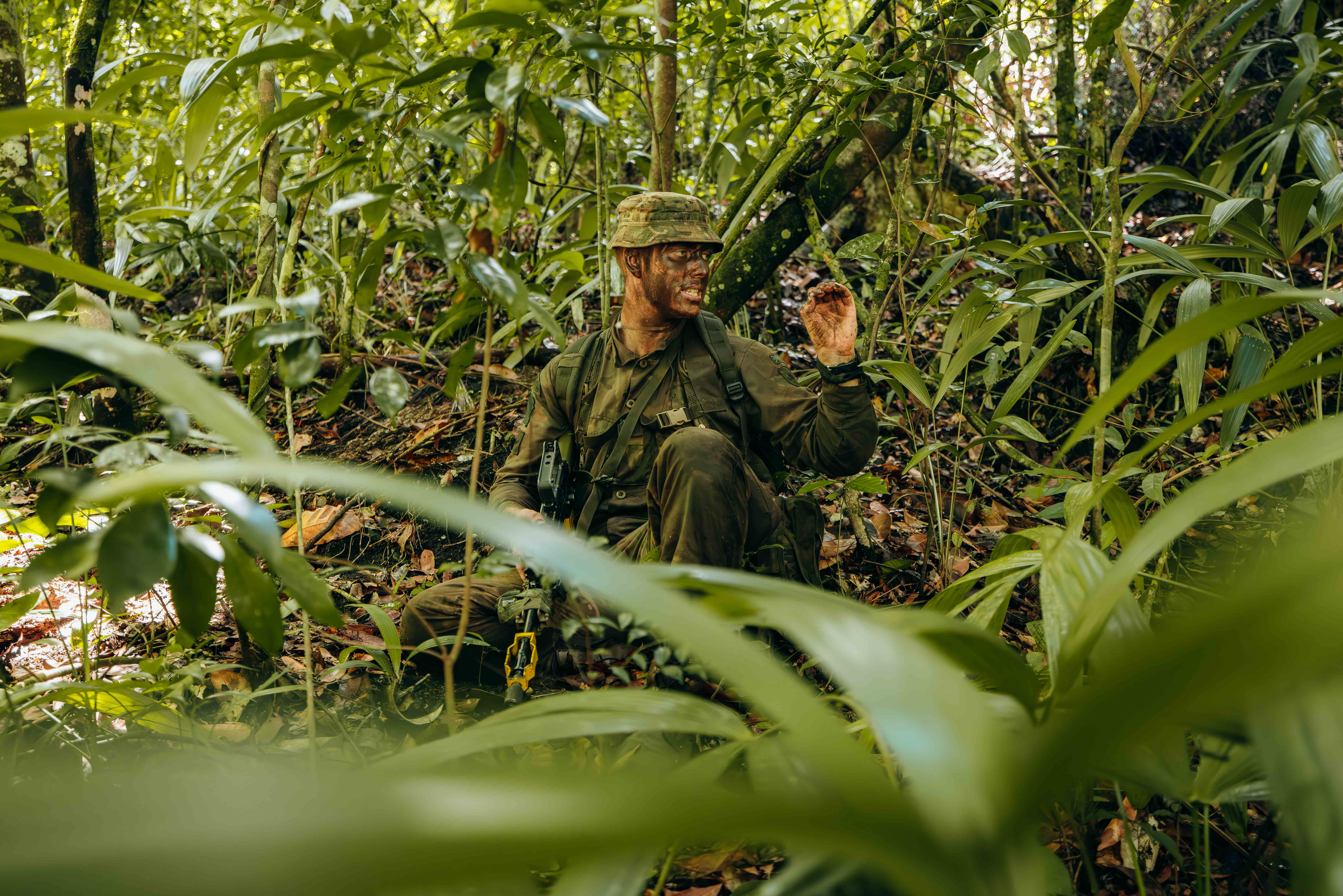 Soldier in camouflage gear crouching in dense jungle foliage.
