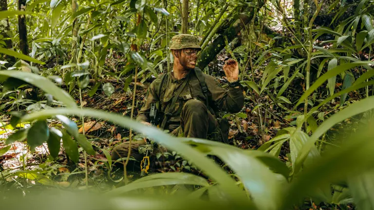 Soldier in camouflage gear crouching in dense jungle foliage.