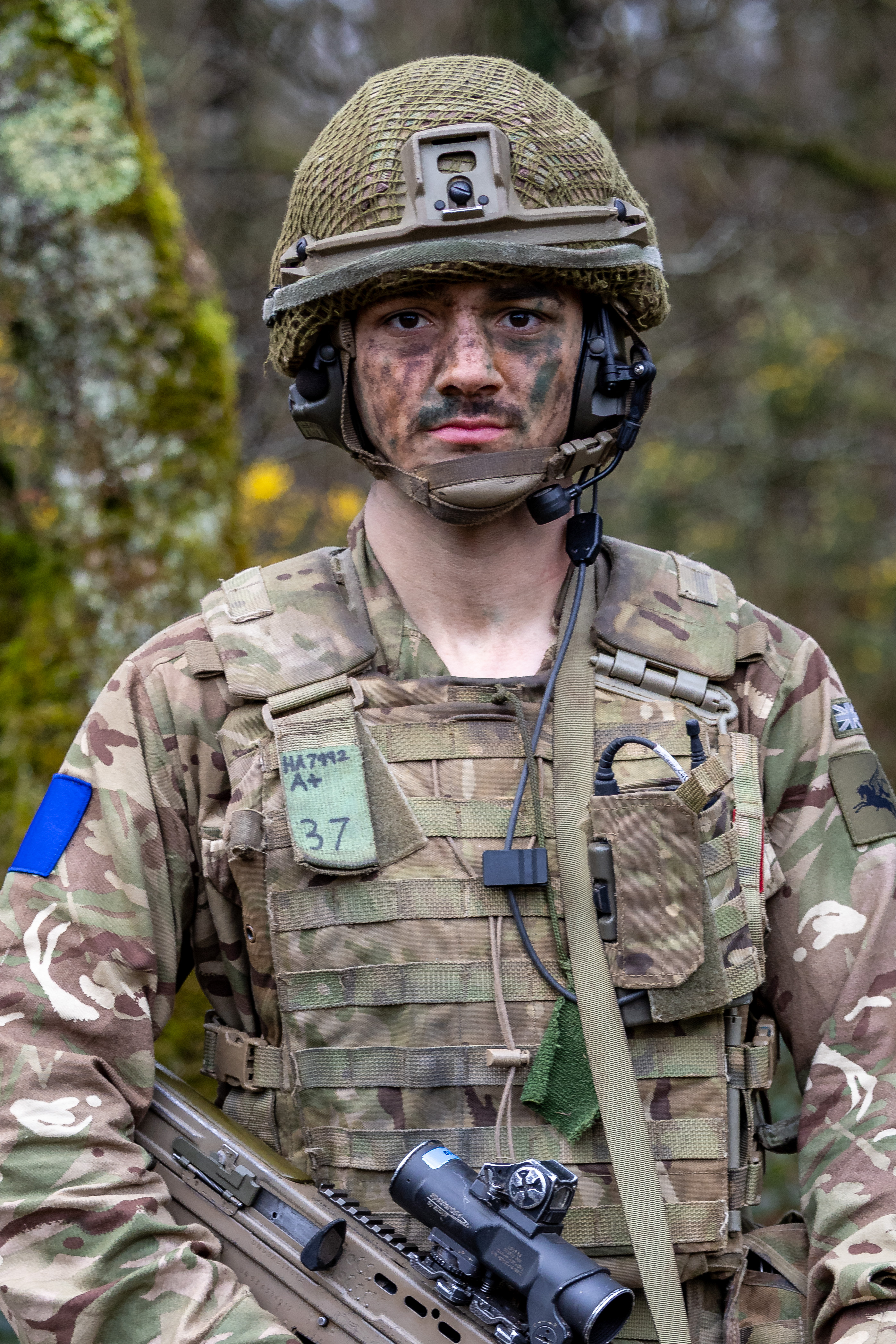A person in military camouflage, wearing a helmet with netting and a communication headset, stands outdoors. 