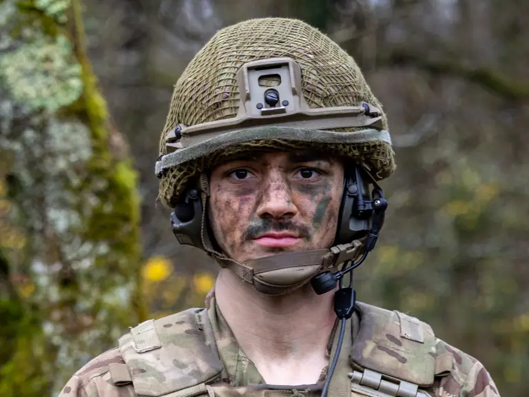 A person in military camouflage, wearing a helmet with netting and a communication headset, stands outdoors.