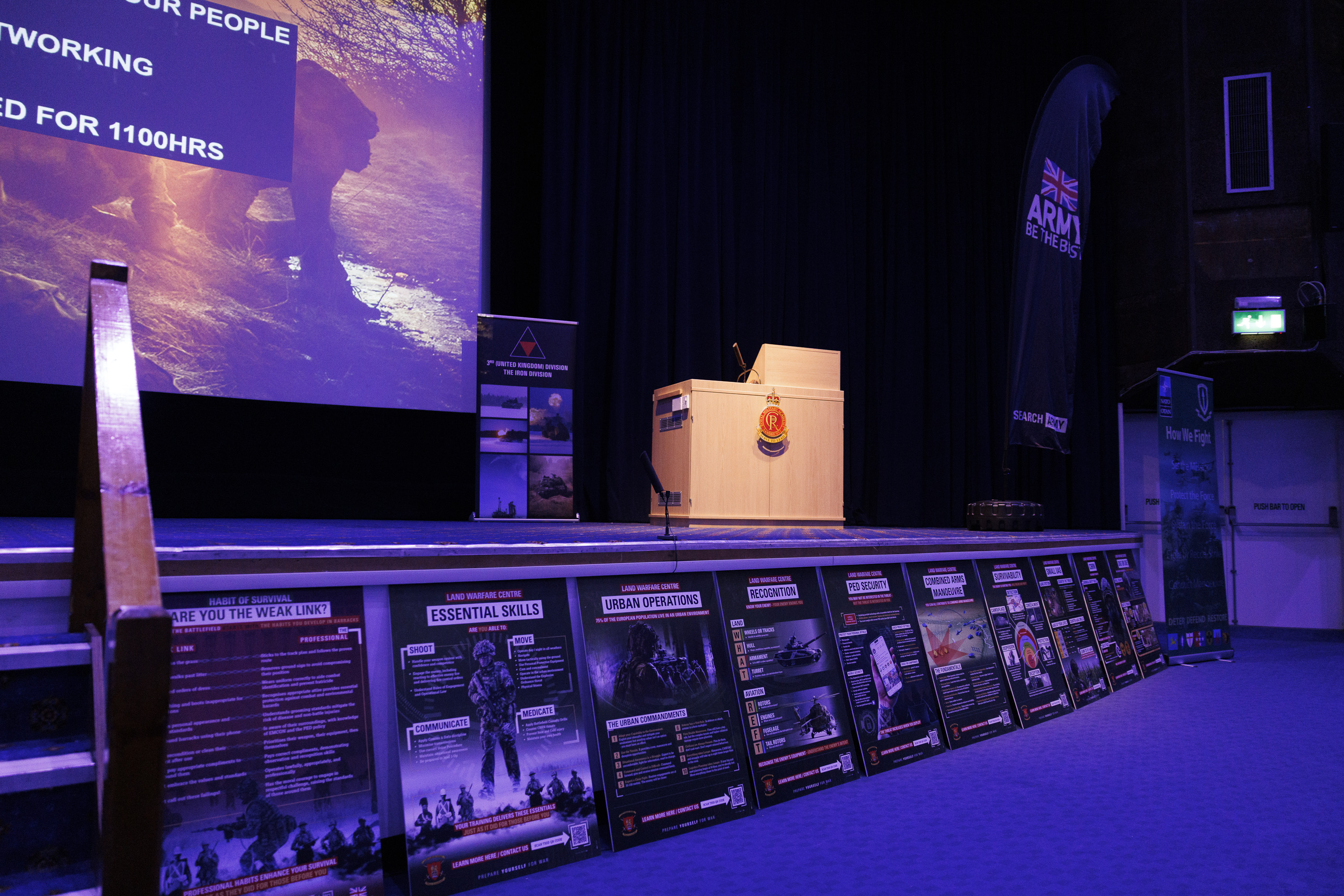 Stage setup for a military presentation with a podium, banners, and posters. A screen displays text. The atmosphere is formal and professional.