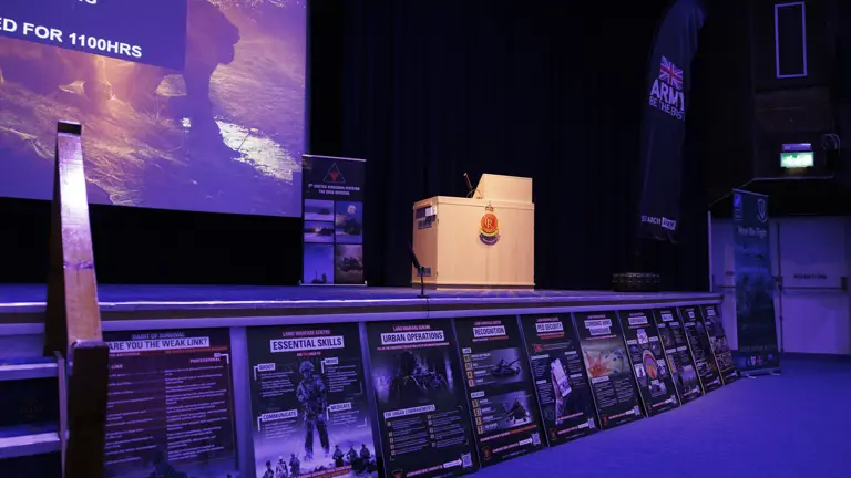 Stage setup for a military presentation with a podium, banners, and posters. A screen displays text. The atmosphere is formal and professional.