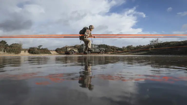 Soldier in full camouflage gear crosses a body of water during a training exercise.