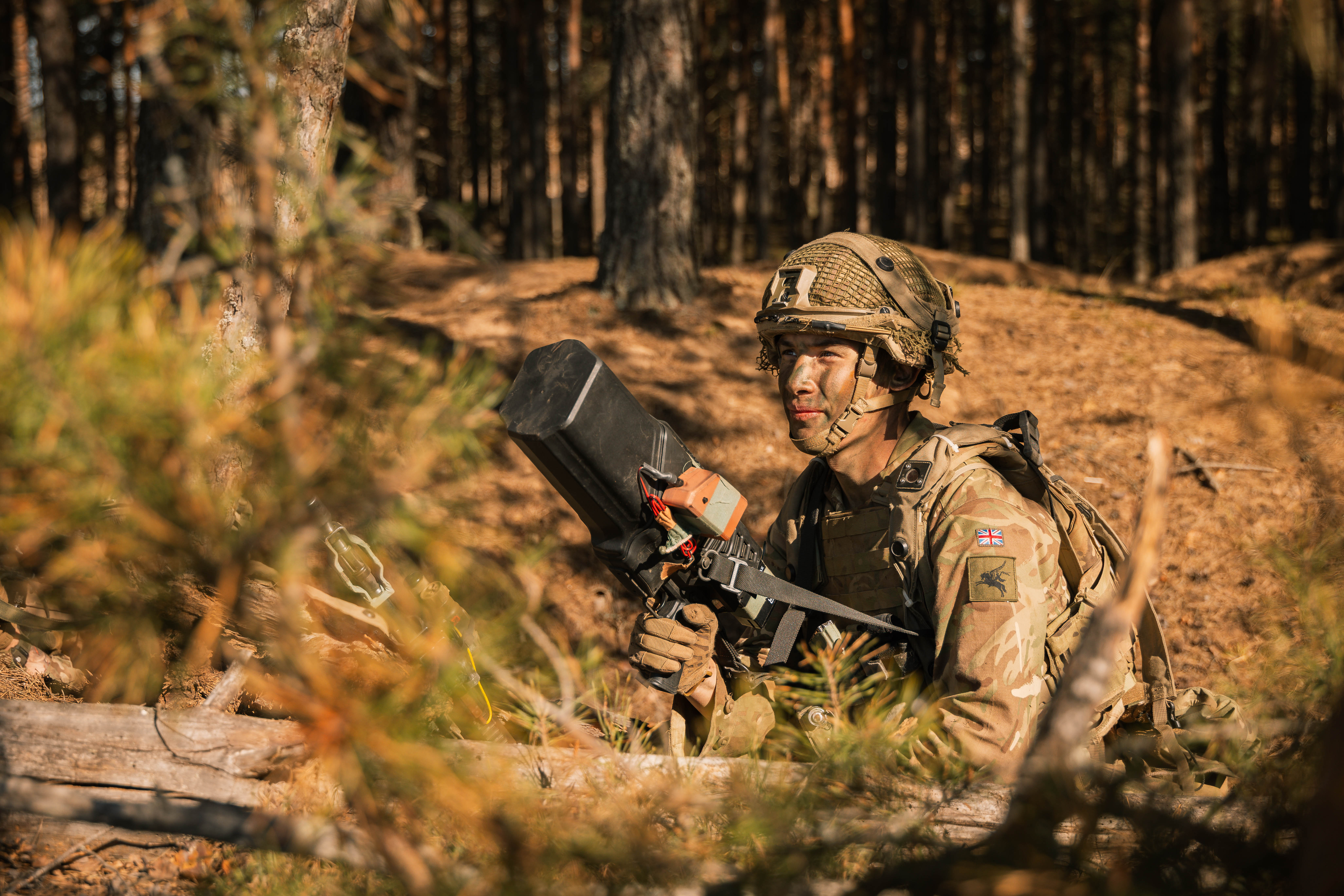 Soldier in camo gear crouches in a forest, holding a black device. Focused expression, dappled sunlight, conveying alertness and readiness.