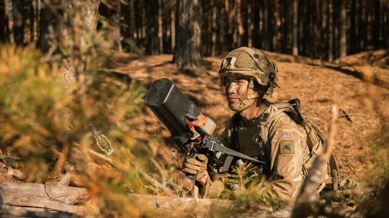 Soldier in camo gear crouches in a forest, holding a black device. Focused expression, dappled sunlight, conveying alertness and readiness.