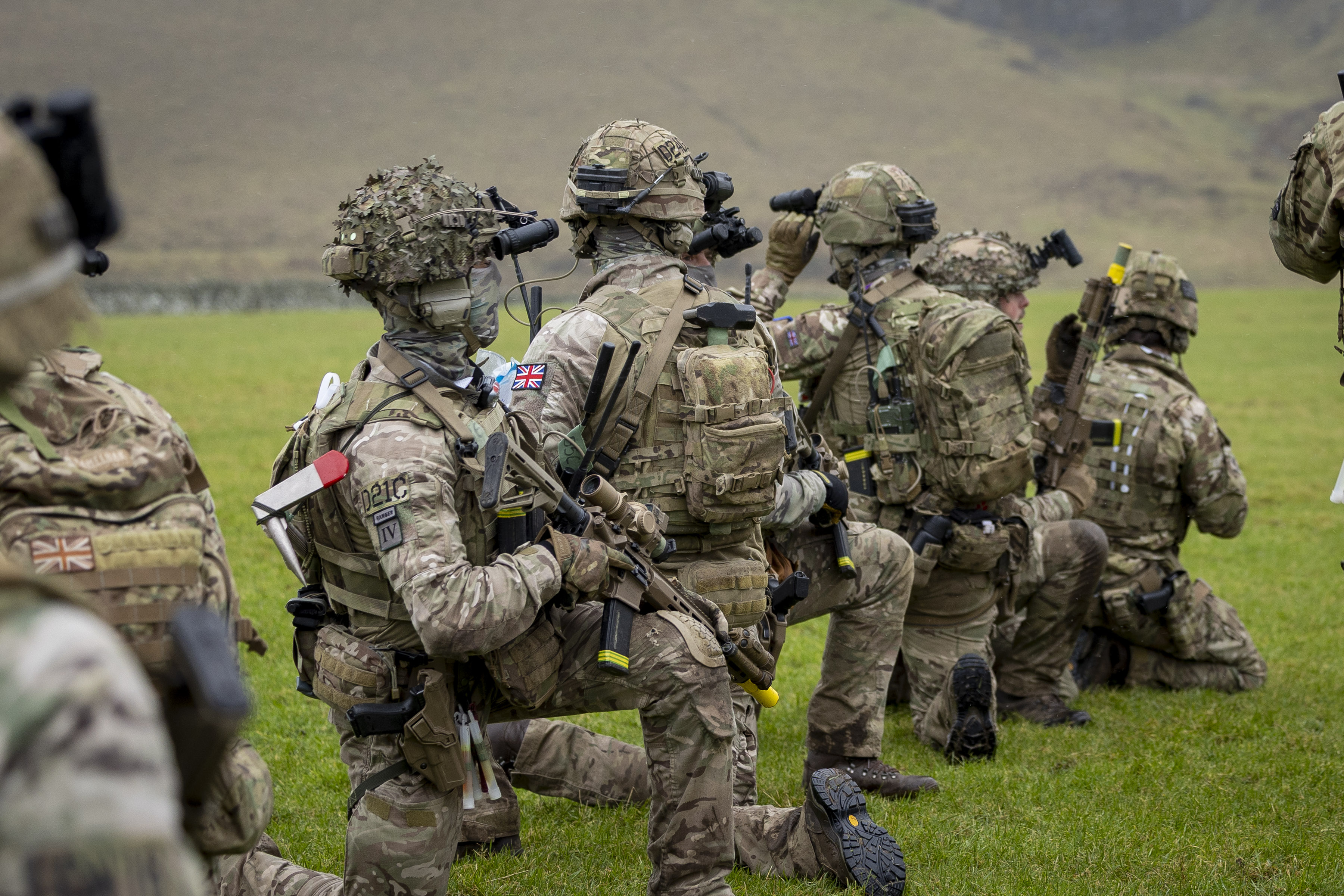 British soldiers in full combat gear kneeling on grass during a training exercise in a rural area.