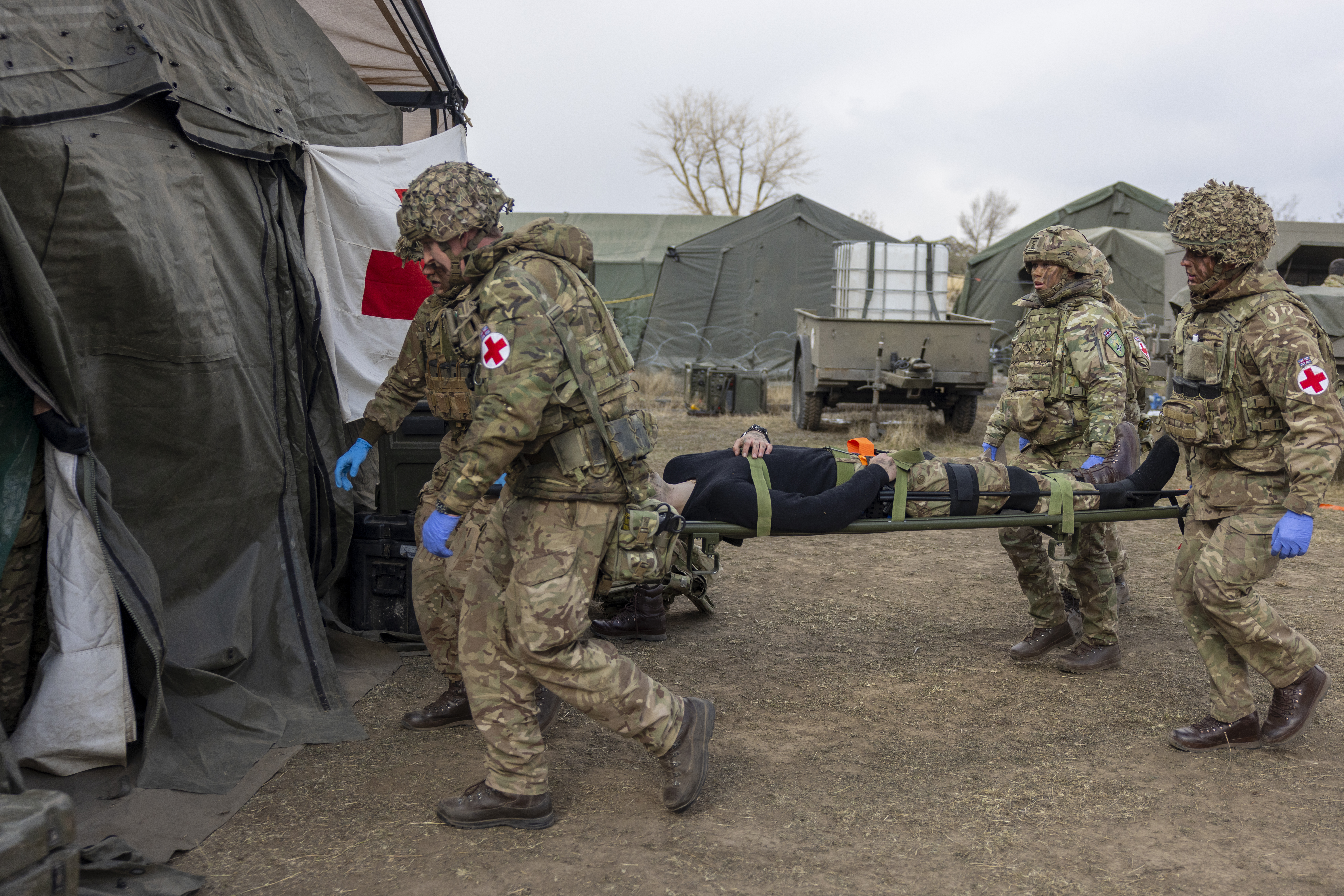 A group of individuals in camouflage uniforms and helmets carry a person on a stretcher towards a medical tent.