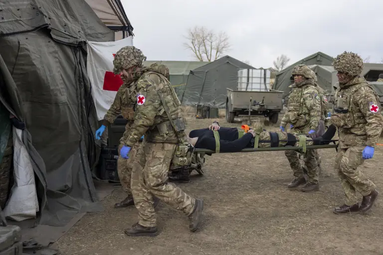A group of individuals in camouflage uniforms and helmets carry a person on a stretcher towards a medical tent.