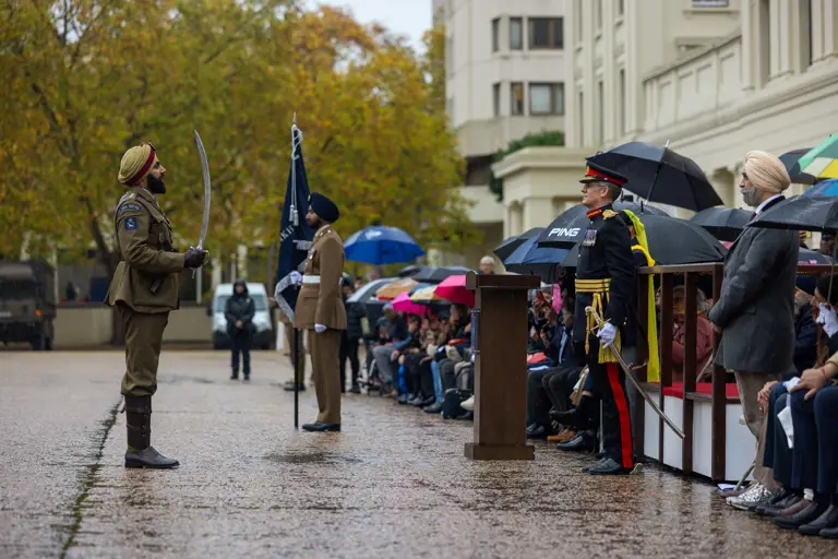 Soldier in traditional uniform holding a sword stands at attention during a formal outdoor ceremony.