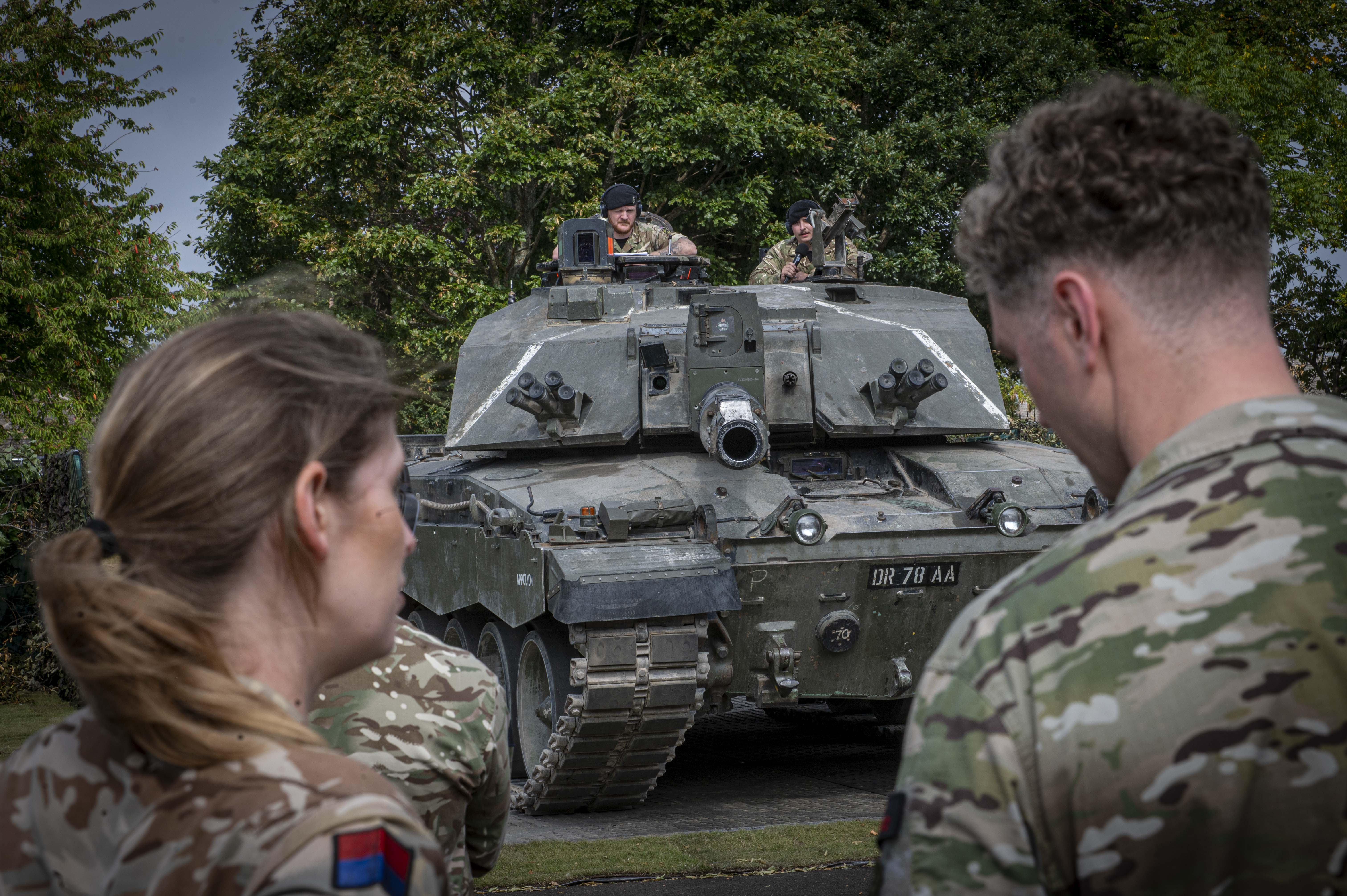 British soldiers observe a Challenger 2 tank parked on a paved area surrounded by trees.