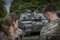 British soldiers observe a Challenger 2 tank parked on a paved area surrounded by trees.