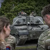 British soldiers observe a Challenger 2 tank parked on a paved area surrounded by trees.
