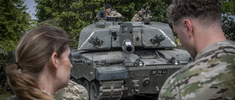 British soldiers observe a Challenger 2 tank parked on a paved area surrounded by trees.