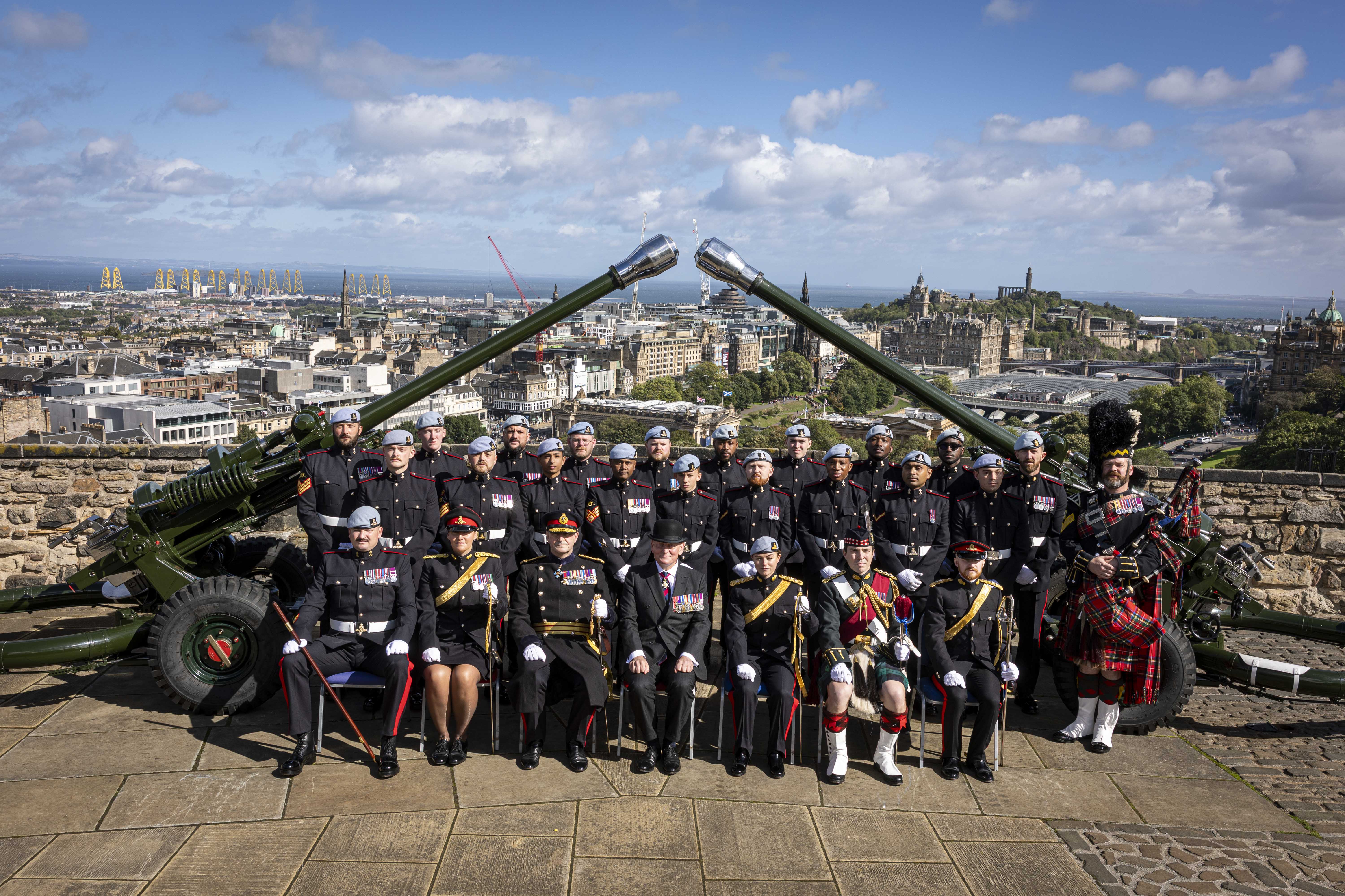 A group of uniformed individuals are posed in front of two large cannons. They are at Edinburgh Castle with a scenic view of the city and the coastline in the background.