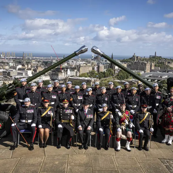A group of uniformed individuals are posed in front of two large cannons. They are at Edinburgh Castle with a scenic view of the city and the coastline in the background.