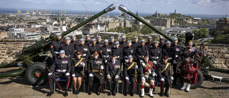 A group of uniformed individuals are posed in front of two large cannons. They are at Edinburgh Castle with a scenic view of the city and the coastline in the background.