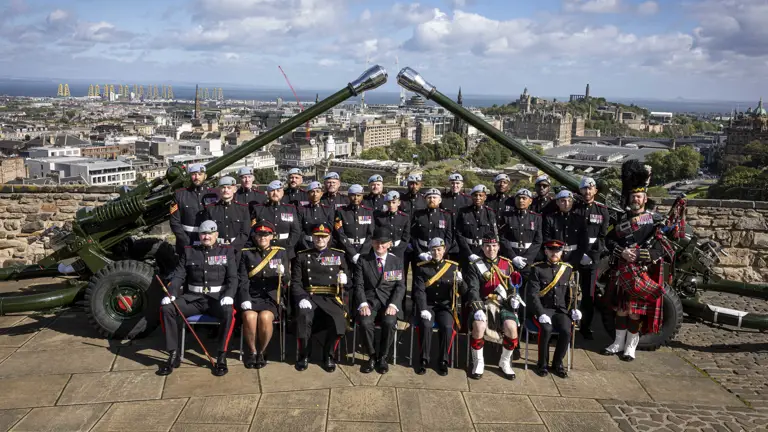 A group of uniformed individuals are posed in front of two large cannons. They are at Edinburgh Castle with a scenic view of the city and the coastline in the background.