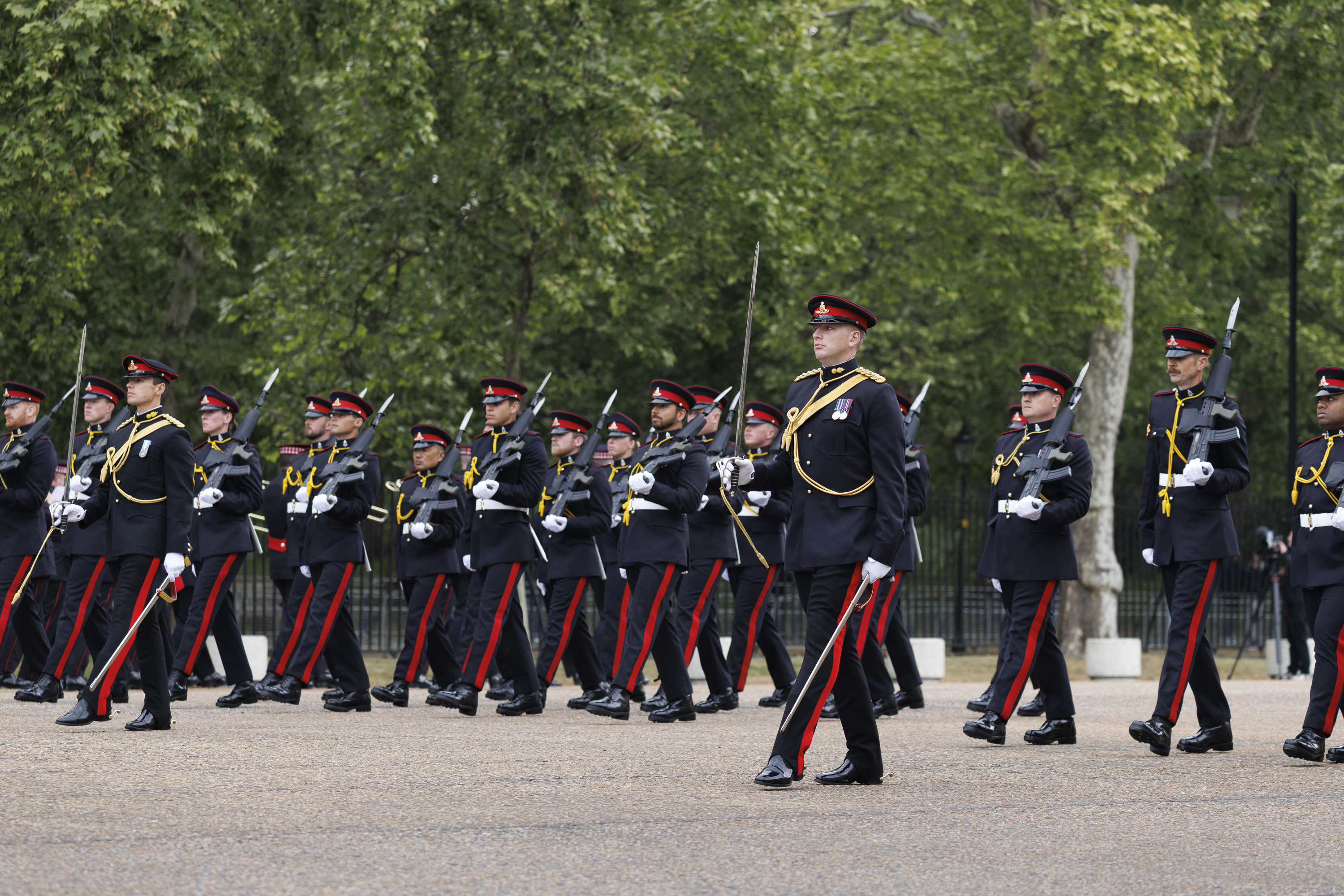 A group of uniformed guards are marching in formation. The guards are wearing dark uniforms with red stripes and caps.