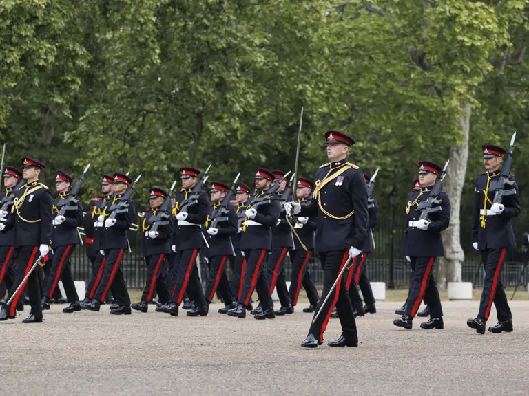A group of uniformed guards are marching in formation. The guards are wearing dark uniforms with red stripes and caps.