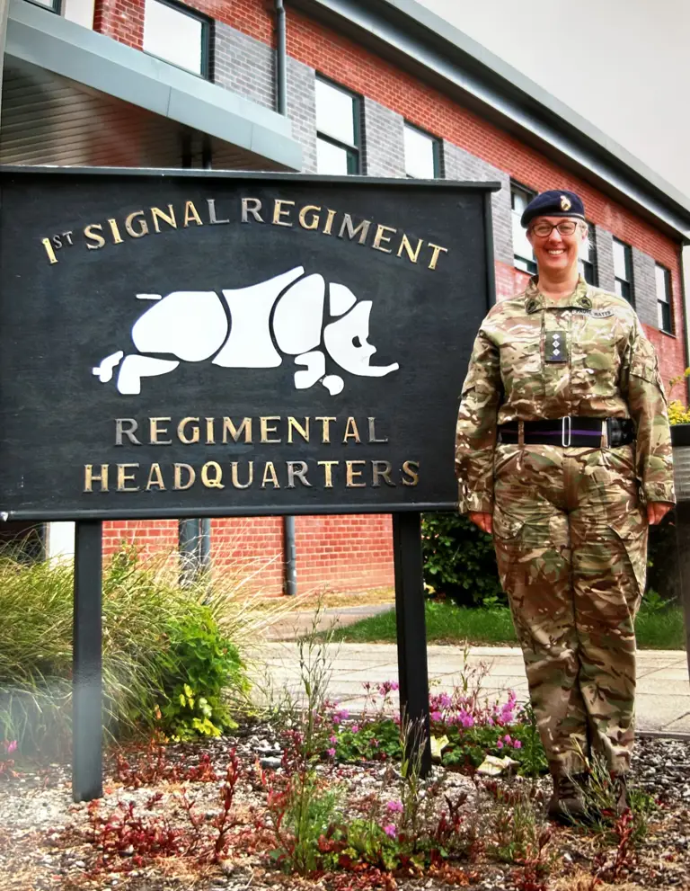 Reverend Lorraine Watts, posing in front of 1st signal regiment, regimental headquarters sign wearing camouflage uniform.