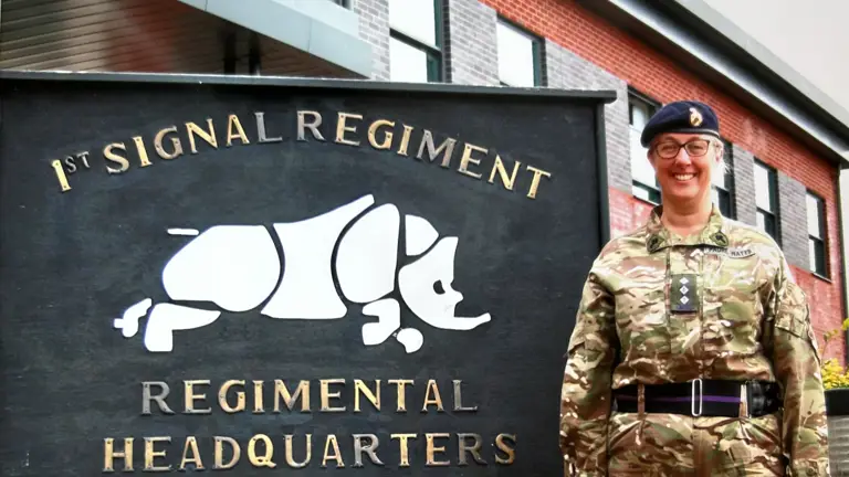 Reverend Lorraine Watts, posing in front of 1st signal regiment, regimental headquarters sign wearing camouflage uniform.