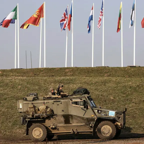 A Foxhound vehicle is shown driving across a road stocked with kit and operated by soldiers. Behind the vehicle is a small hill where onto shows the flags of ten various different NATO countries.