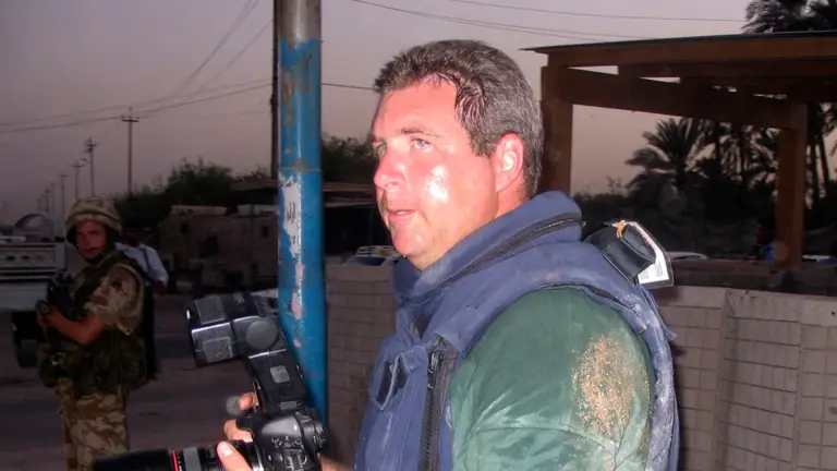 Soldier magazine journalist seen with a camera wearing a bullet proof vest while on a job in Iraq, a soldier can be seen in the background.