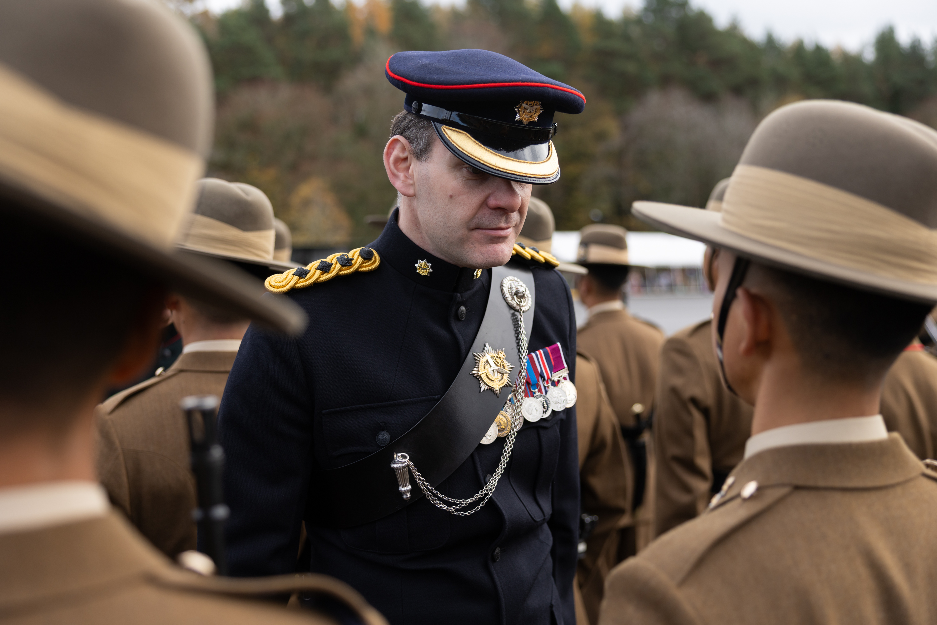 A uniformed officer with medals and a peaked cap converses with soldiers in brown uniforms and hats, set against a backdrop of trees.