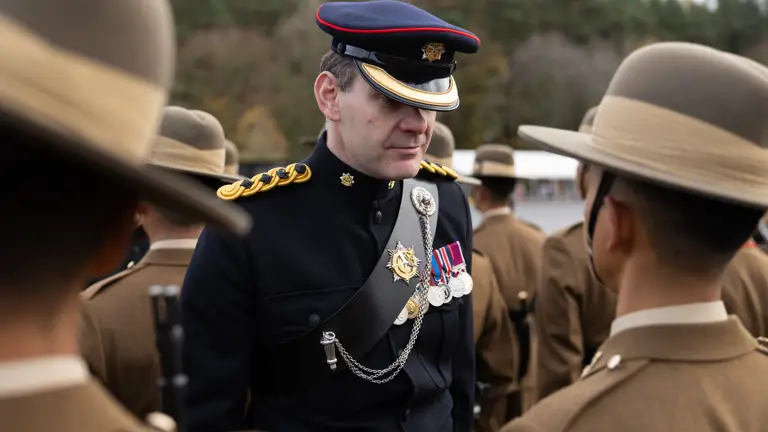 A uniformed officer with medals and a peaked cap converses with soldiers in brown uniforms and hats, set against a backdrop of trees.