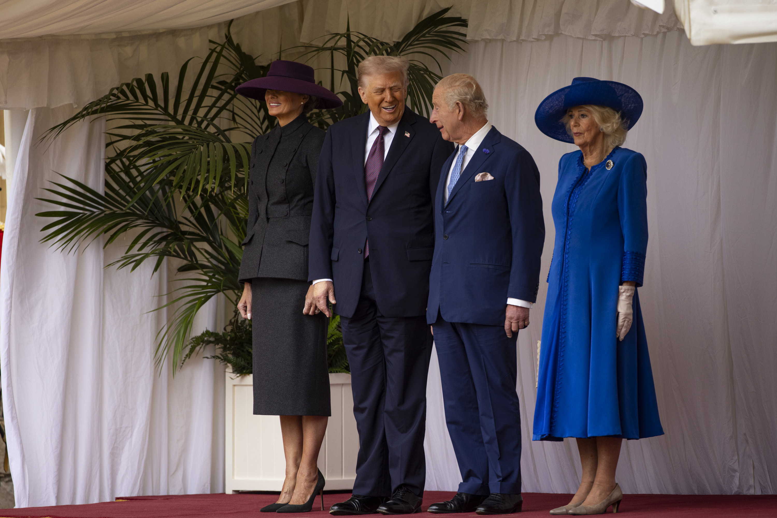 Two men in dark suits and two women in formal dresses and hats stand on a red carpeted stage with white curtains.
