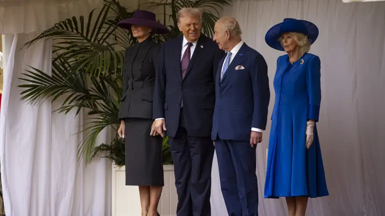 Two men in dark suits and two women in formal dresses and hats stand on a red carpeted stage with white curtains.