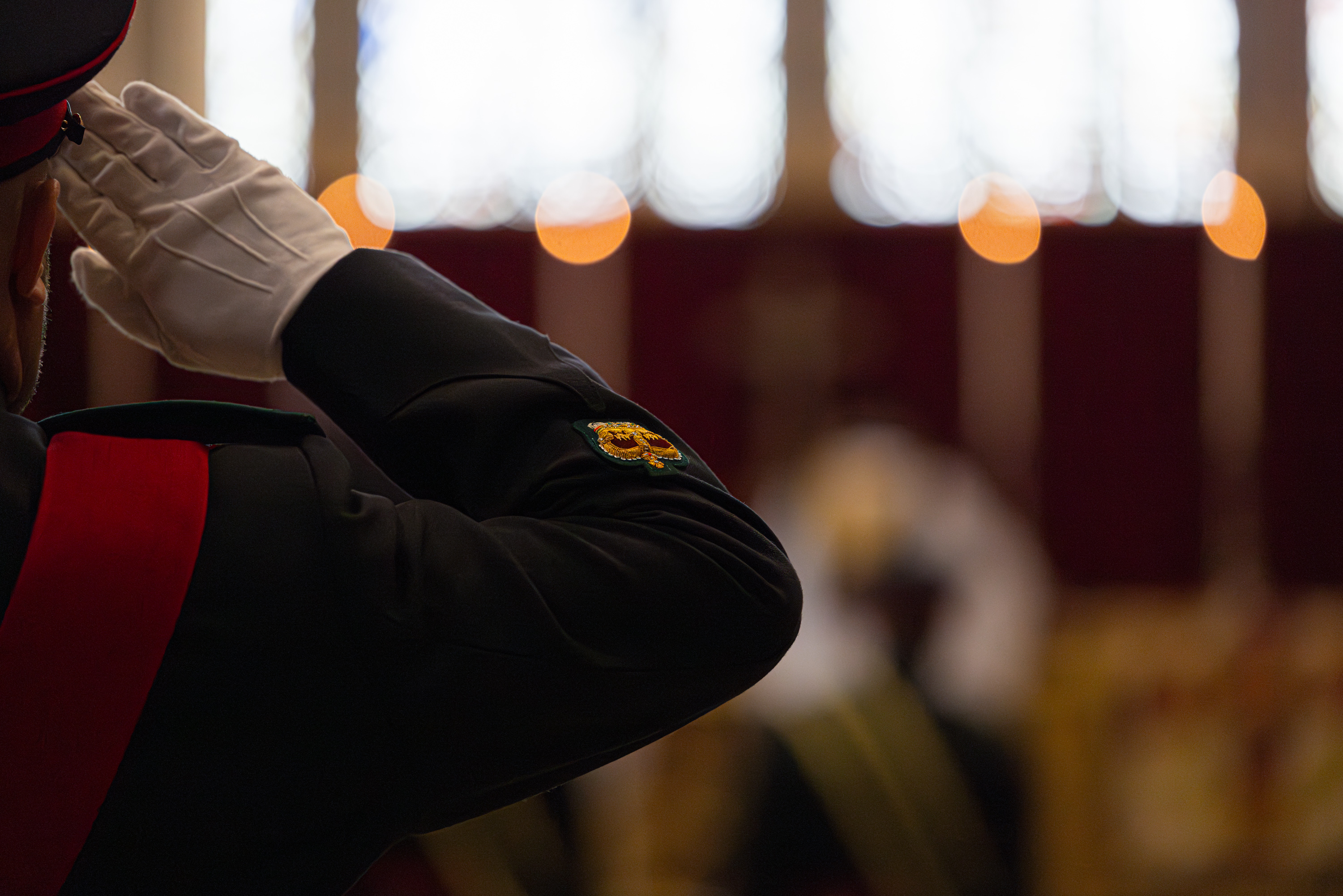 A person in a formal military uniform is saluting in the foreground. The person is wearing white gloves and a ceremonial jacket with a distinctive patch. In the background, the scene is blurred, showing an indoor setting with subtle lighting, suggesting a formal or ceremonial event.