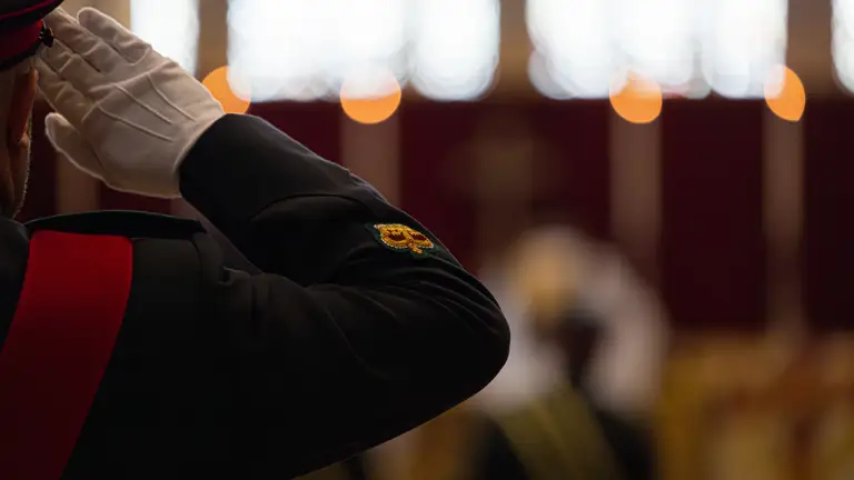 A person in a formal military uniform is saluting in the foreground. The person is wearing white gloves and a ceremonial jacket with a distinctive patch. In the background, the scene is blurred, showing an indoor setting with subtle lighting, suggesting a formal or ceremonial event.