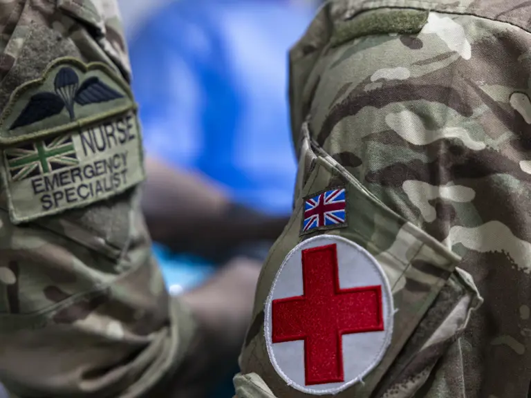 Image shows a close up of two soldiers arms next to each other. The right arm has a flash that says 'Nurse, Emergency Specialist' and the left arm has a Union Jack Flag and a red cross flash.