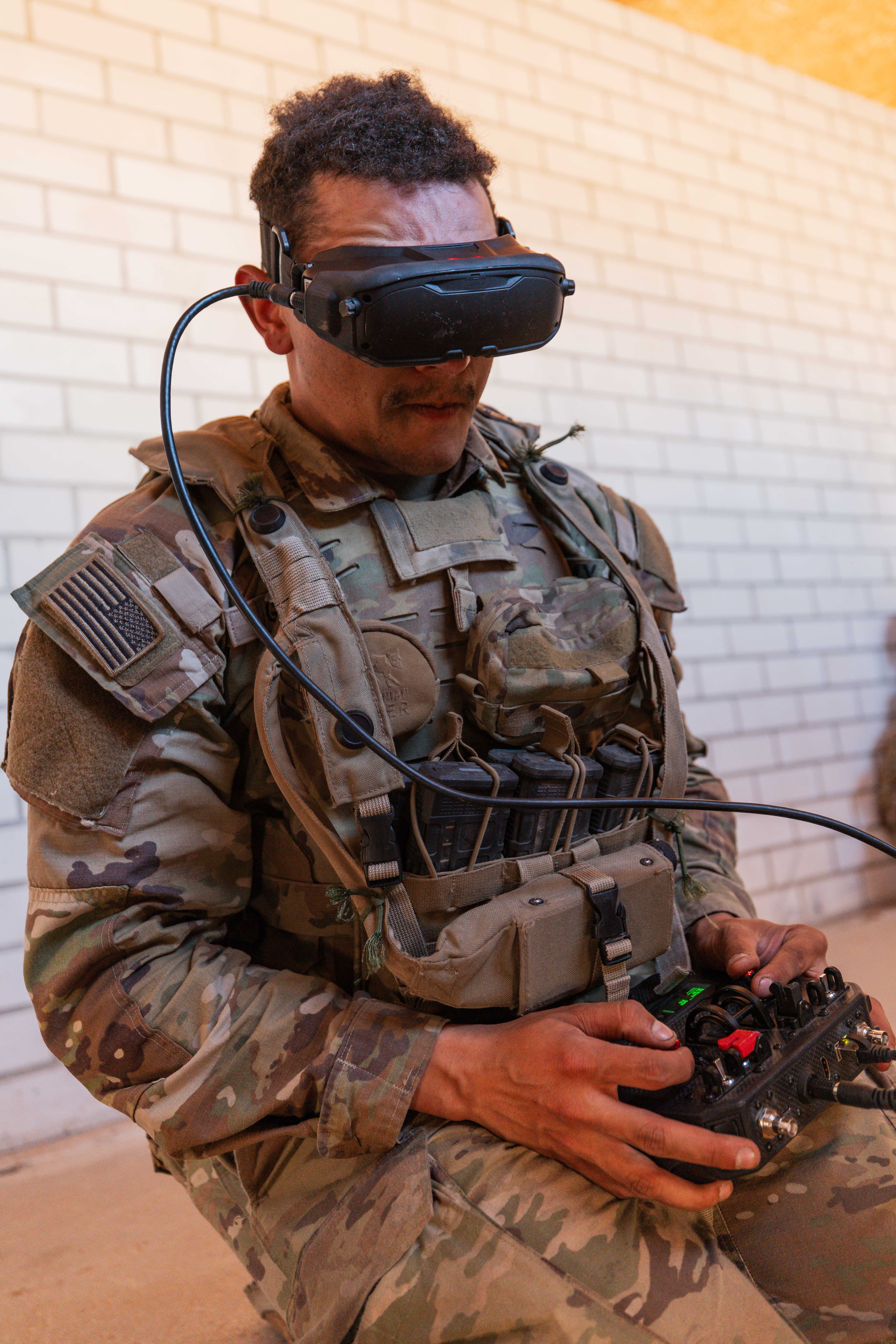 A soldier in camouflage gear operates a remote control while wearing VR goggles, focused and intent. The setting is a room with light brick walls.