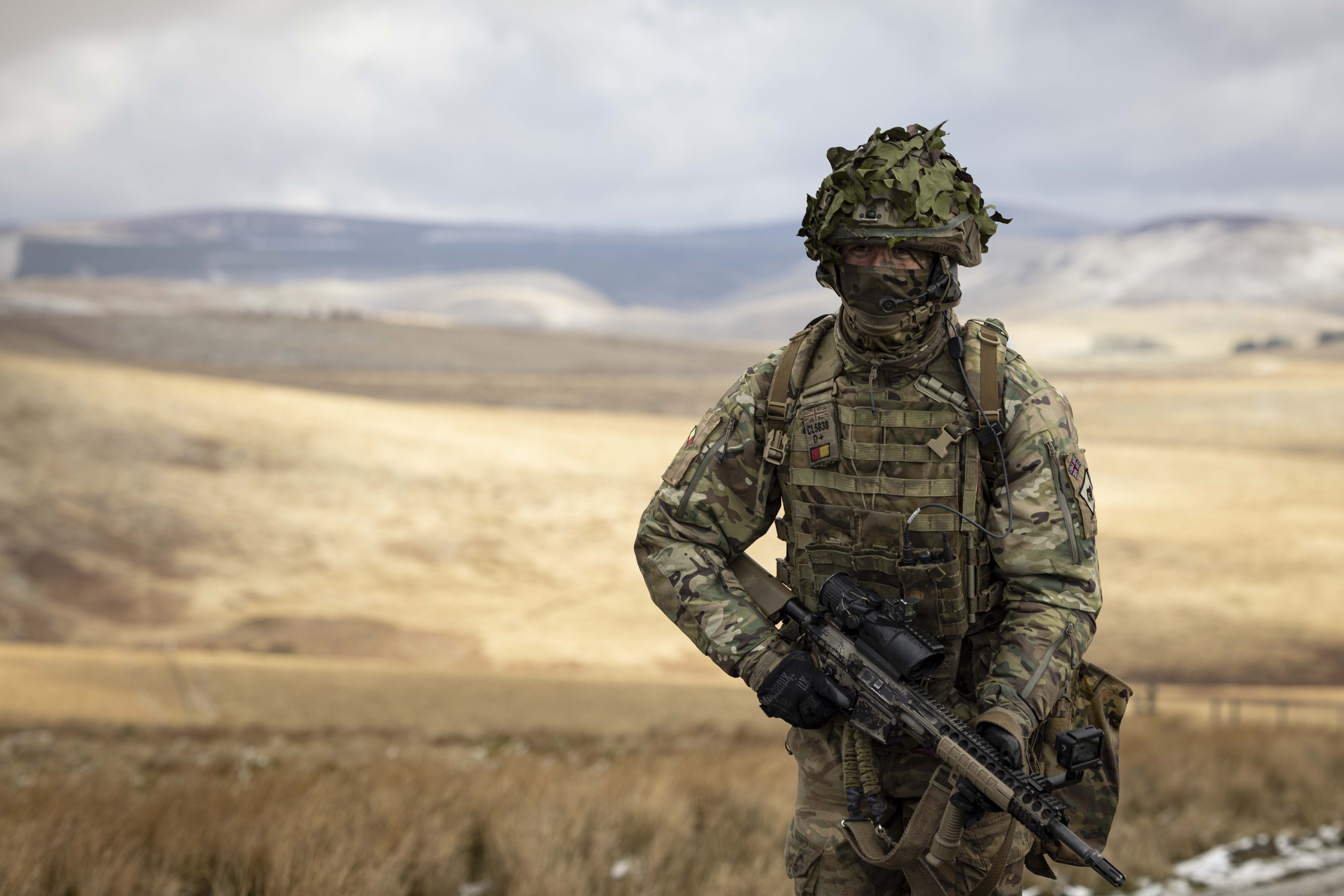 Soldier in camouflage uniform wearing body armour and helmet walks in the firing range holding his rifle.