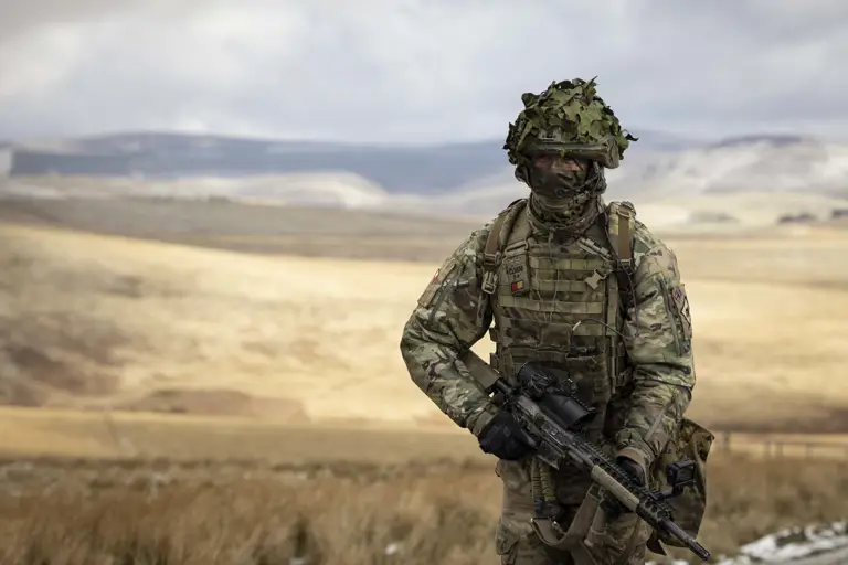 Soldier in camouflage uniform wearing body armour and helmet walks in the firing range holding his rifle.