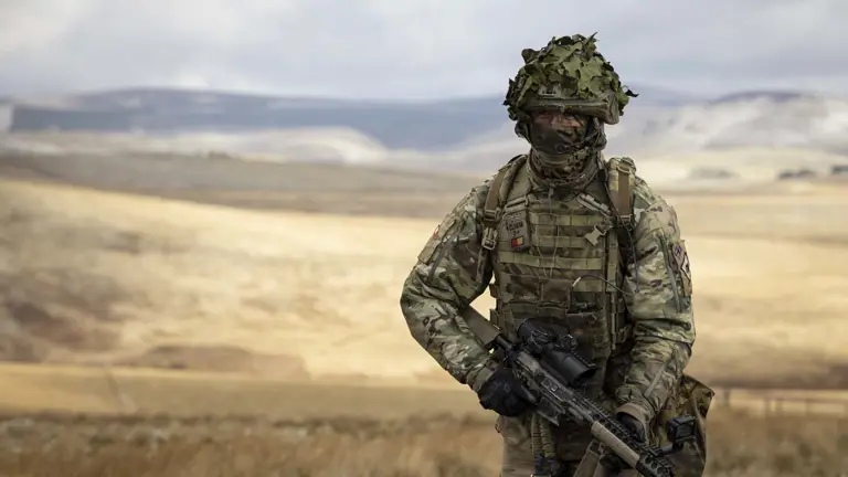 Soldier in camouflage uniform wearing body armour and helmet walks in the firing range holding his rifle.