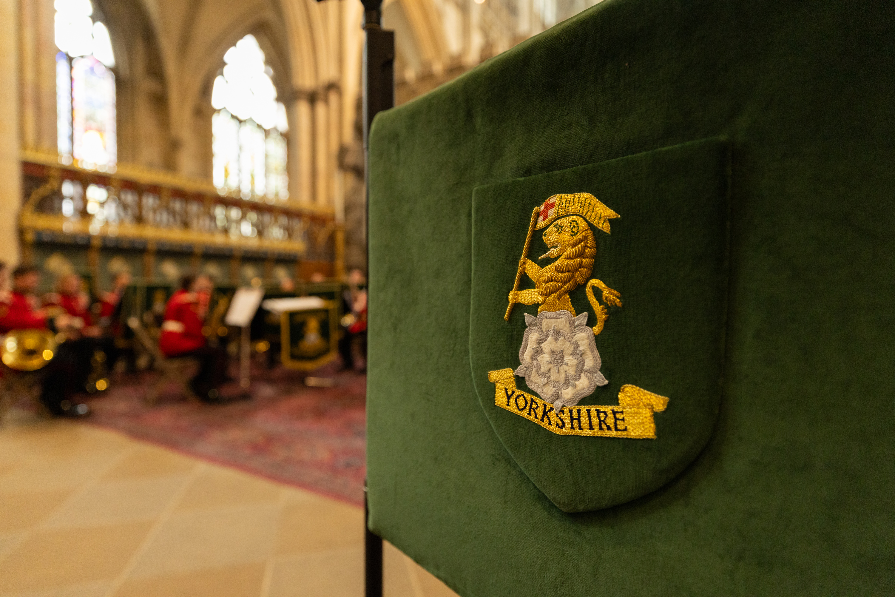 A green embroidered banner with the Yorkshire emblem is in the foreground. In the background, there is a blurred view of a musical ensemble in red uniforms performing inside a cathedral or large hall. The scene includes ornate architecture and musical stands.