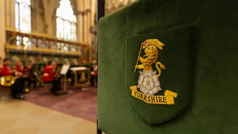 A green embroidered banner with the Yorkshire emblem is in the foreground. In the background, there is a blurred view of a musical ensemble in red uniforms performing inside a cathedral or large hall. The scene includes ornate architecture and musical stands.