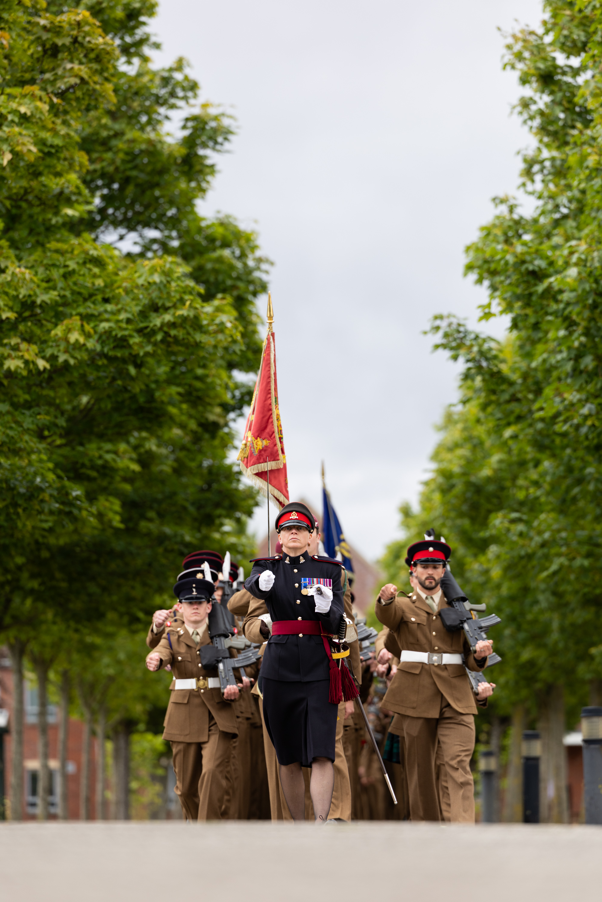 A group of people in military uniforms is marching in formation down a tree-lined street. One person at the front is holding a ceremonial flag, and several others are holding rifles. The scene is outdoors, under a cloudy sky.