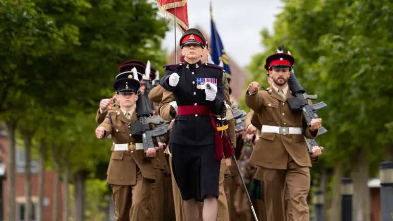 A group of people in military uniforms is marching in formation down a tree-lined street. One person at the front is holding a ceremonial flag, and several others are holding rifles. The scene is outdoors, under a cloudy sky.