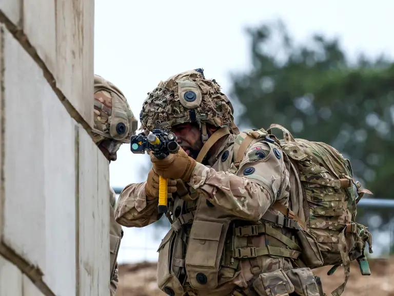 A person in camouflage military gear is aiming a weapon around a corner of a building.