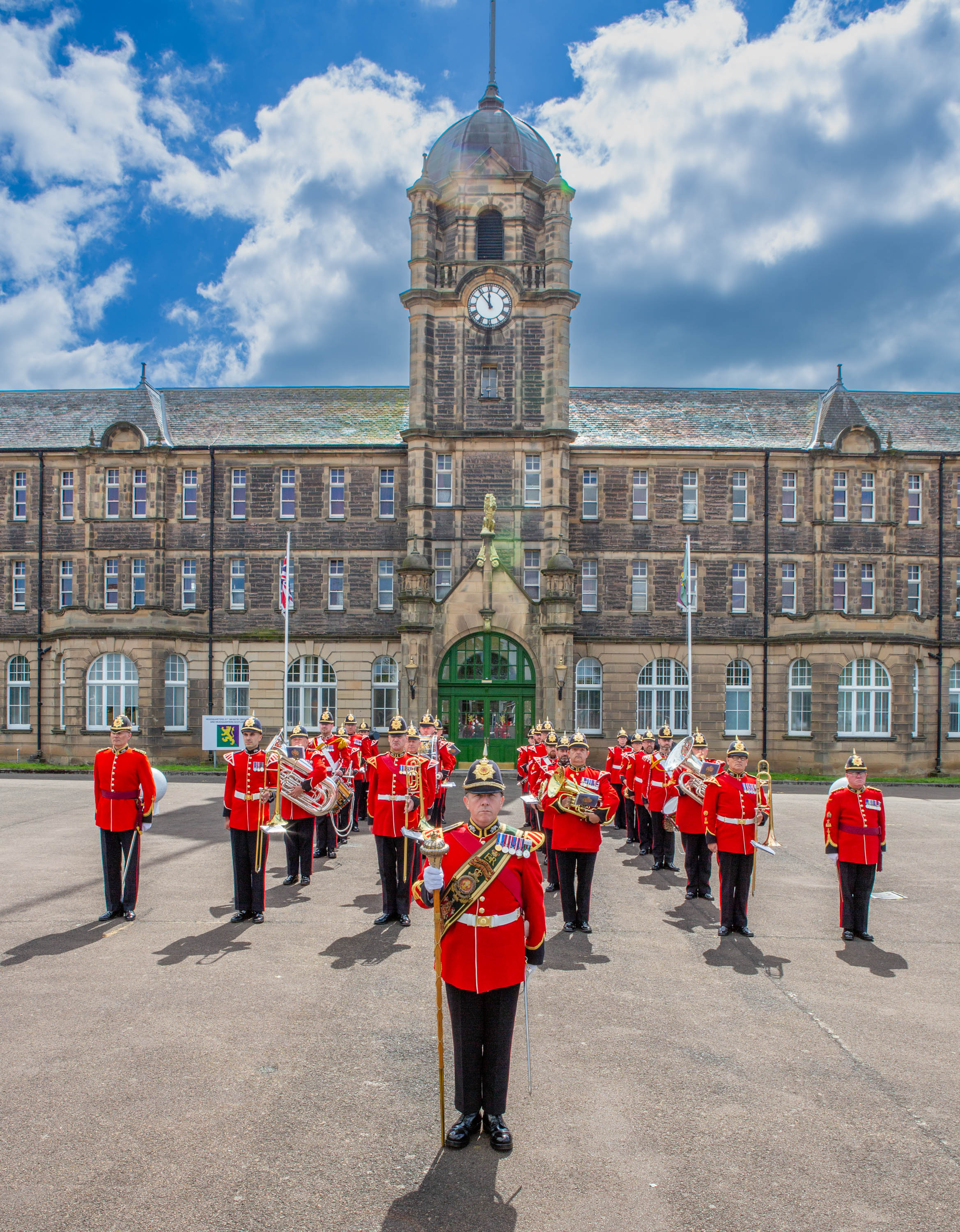 A marching band in bright red uniforms is assembled in formation on a paved area in front of an ornate, historic building.