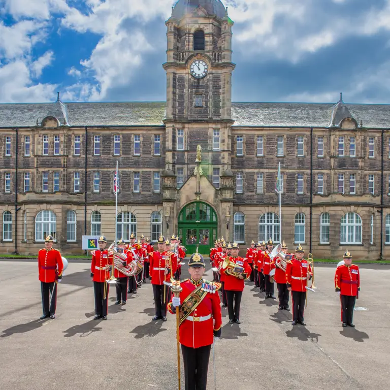 A marching band in bright red uniforms is assembled in formation on a paved area in front of an ornate, historic building.
